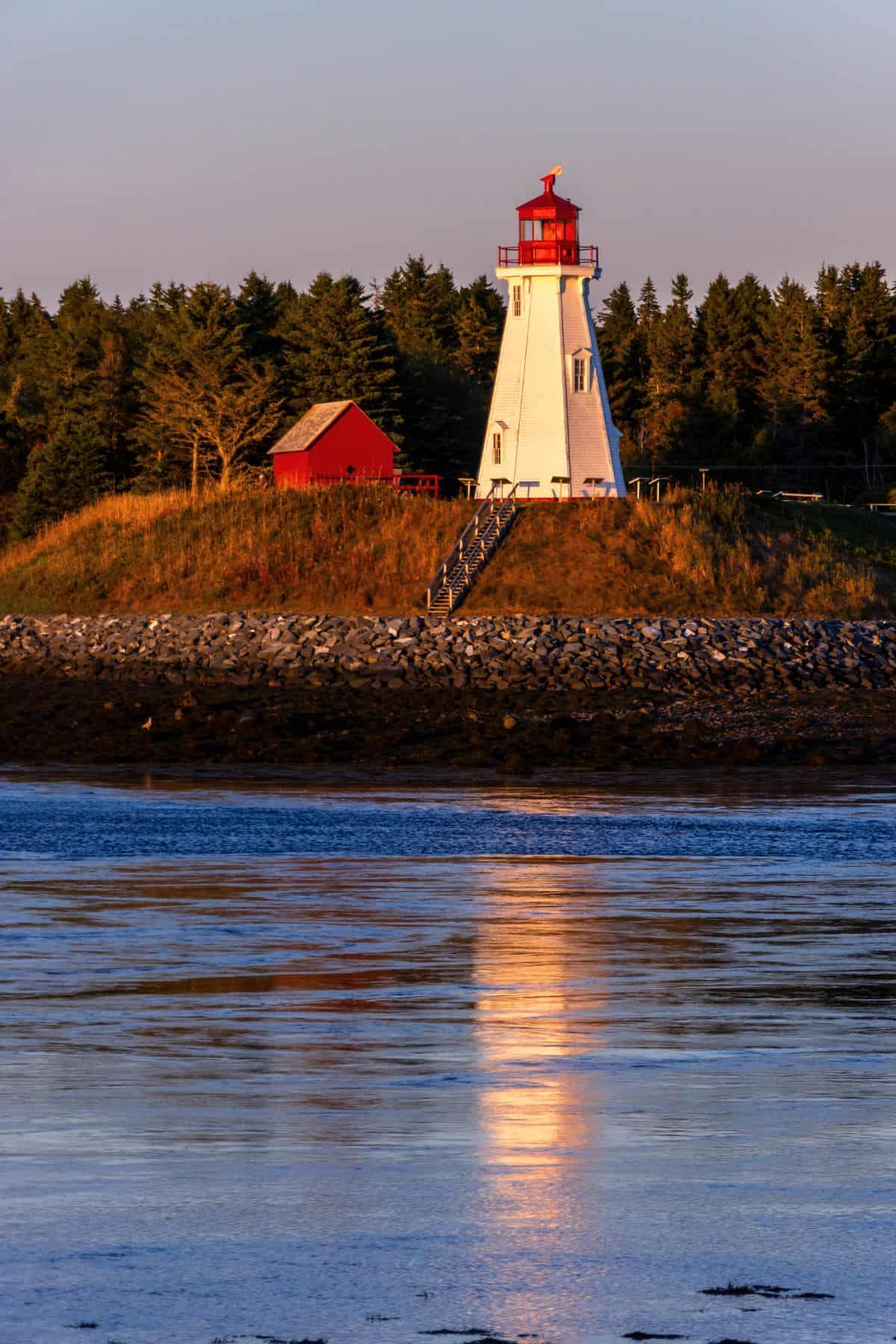 A white lighthouse with a red top stands on a grassy hill near a small red building, with its reflection visible in the calm water below.