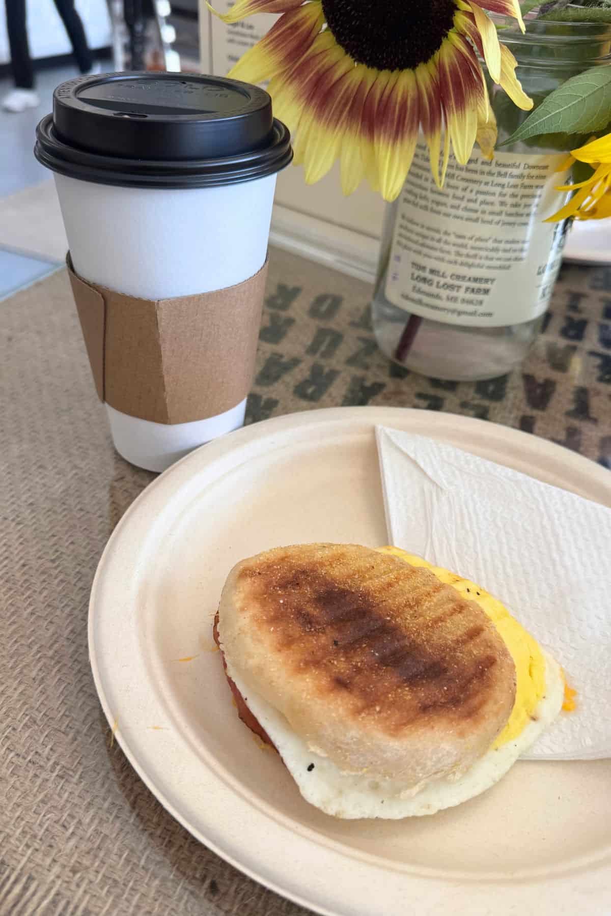 A breakfast sandwich on a paper plate with a folded napkin sits next to a takeaway coffee cup; a vase with a sunflower is in the background.