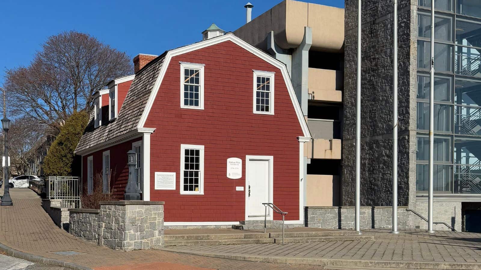 A small red wooden building with white trim stands in front of a concrete parking structure and a glass stairwell on a sunny day.
