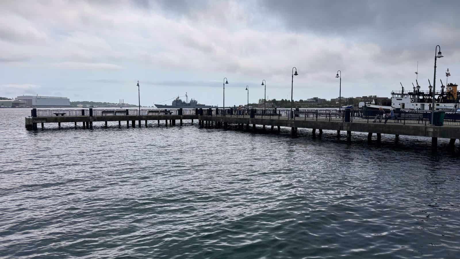 A wooden pier extends over calm water under a cloudy sky, with ships and distant buildings visible in the background.