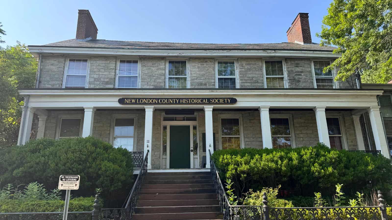 A two-story stone building with a sign reading "New London County Historical Society" above the entrance and a porch surrounded by greenery.
