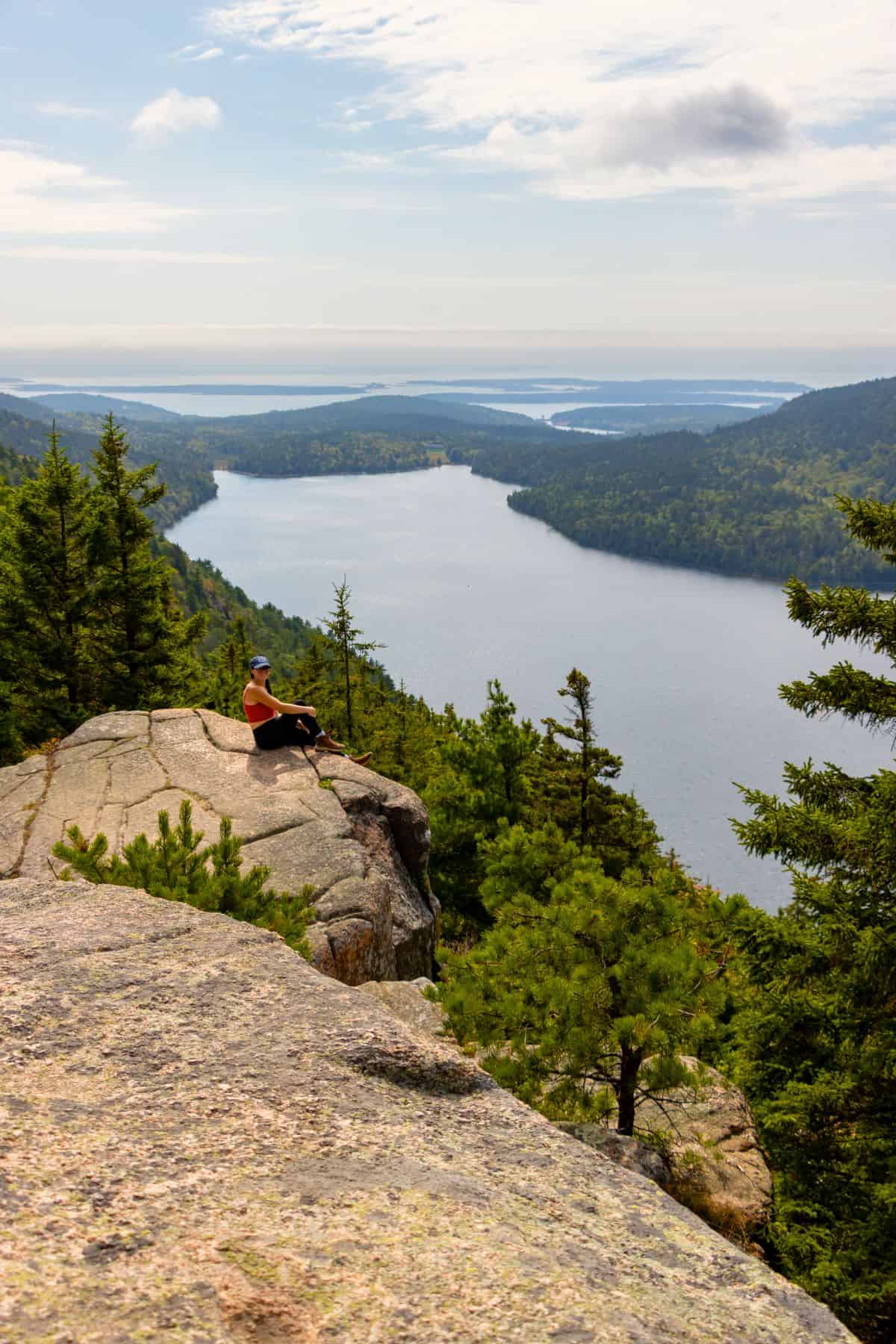 A person sits on a rocky ledge overlooking a forested landscape and a large lake under a partly cloudy sky.