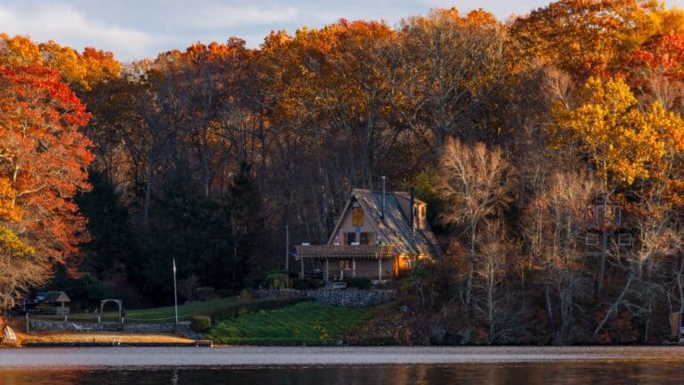 A-frame house surrounded by autumn trees on a lakeshore, with sunlight illuminating the colorful foliage and water in the foreground.