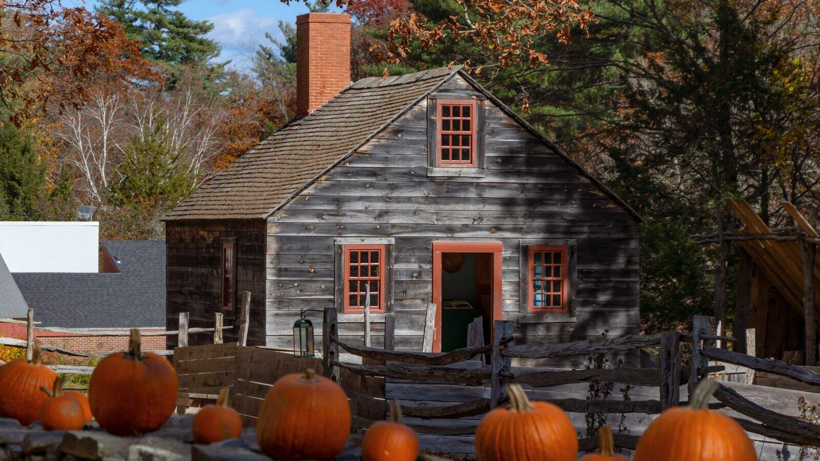 A rustic wooden house with red-trimmed windows and door sits behind a wooden fence lined with pumpkins on an autumn day.