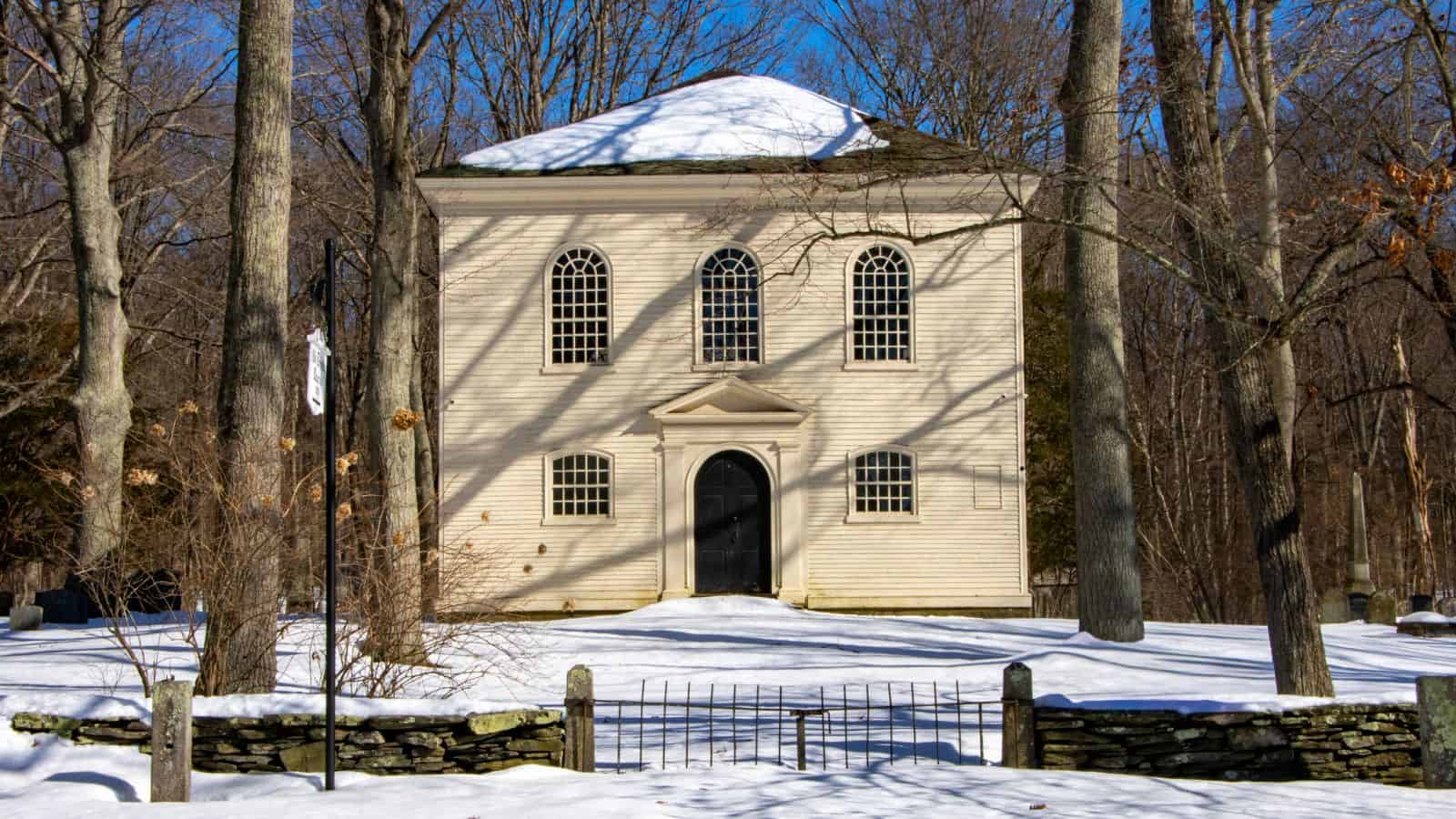 A small, two-story white chapel with arched windows sits among leafless trees and snow, behind a low stone wall and metal gate—one of the rumored haunted places in Connecticut.