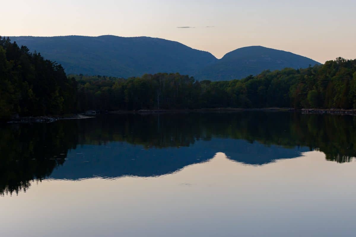 A calm lake reflects a forested shoreline and distant mountains under a clear sky at dusk.