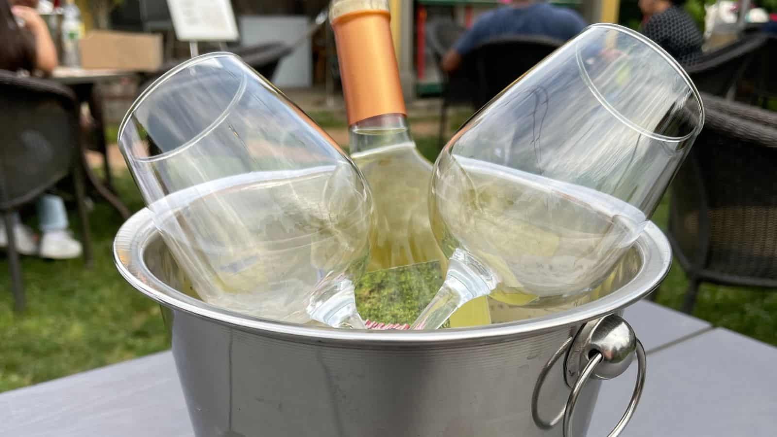A bottle of white wine and two empty wine glasses are placed in an ice bucket on an outdoor table, with people seated in the background.