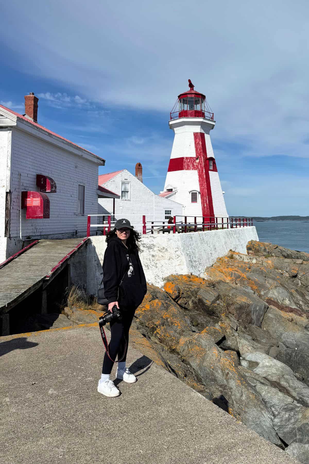 A person in dark clothing stands on a rocky path in front of a white and red lighthouse by the sea near Lubec and Campobello Island on a sunny day.