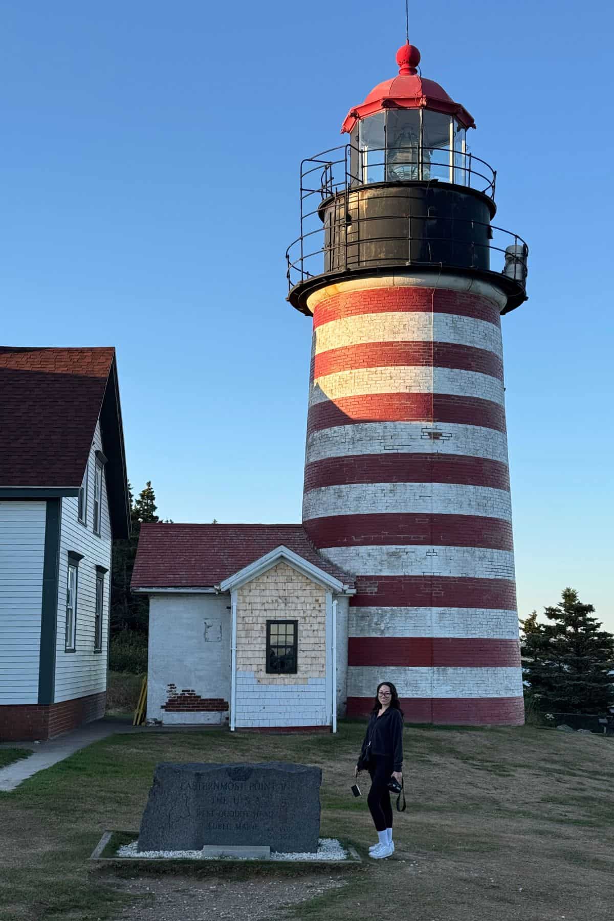 A person stands near a stone monument in front of a red-and-white striped lighthouse with clear skies in the background.