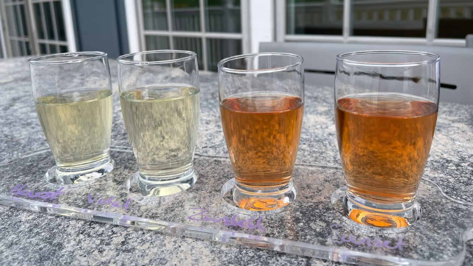 Four glasses of cider in a tasting flight are arranged in a row on a tray, each glass containing a different color and labeled underneath.