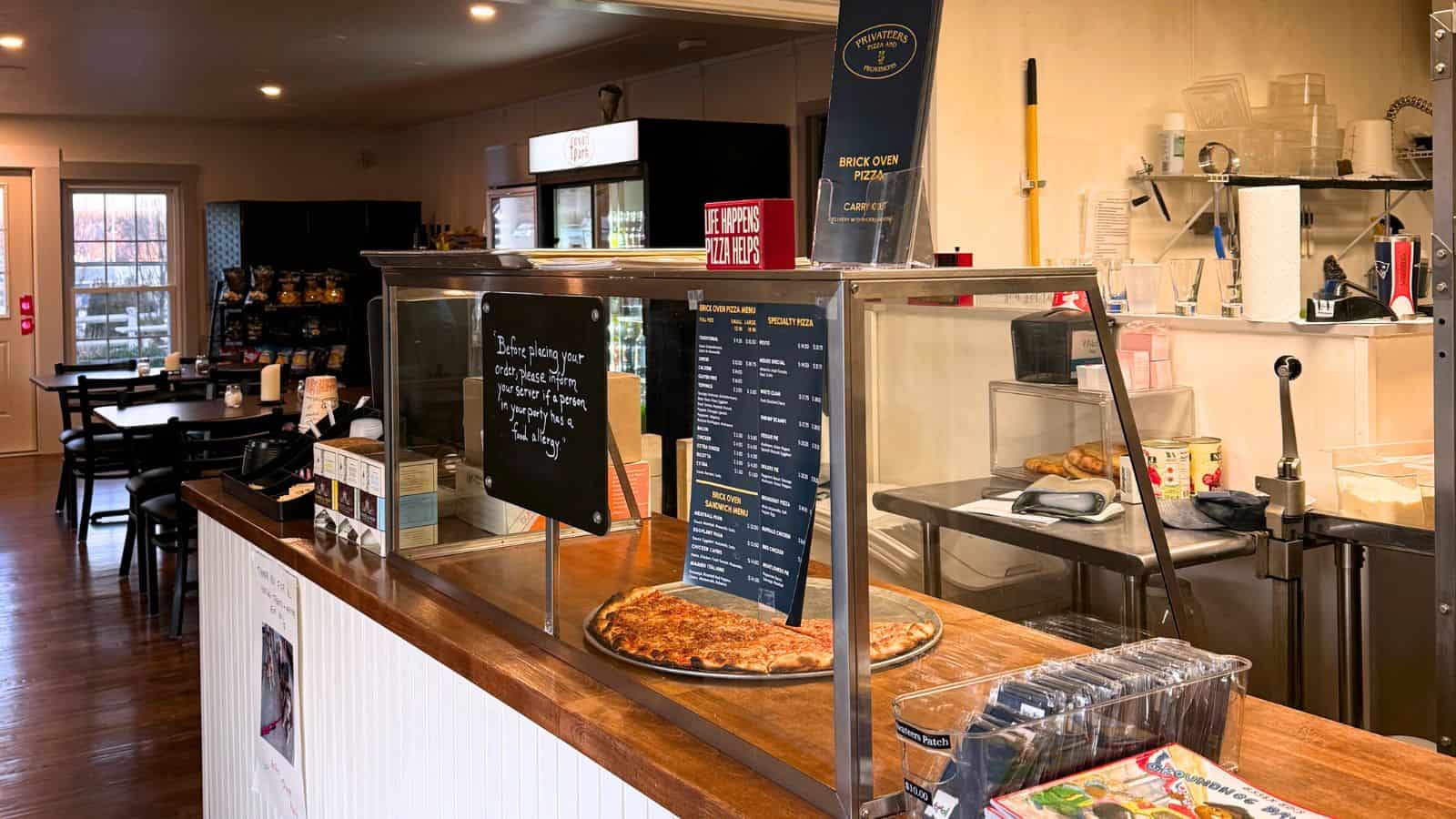 A pizza sits on the counter behind a glass shield at a small café; menus and baked goods are visible, with tables and chairs in the background.