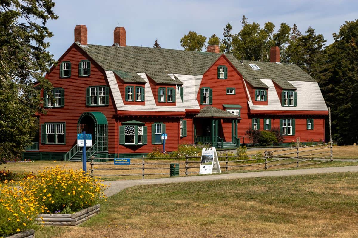 A large red wooden house with green trim sits on a grassy lawn, surrounded by a wooden fence and flower beds, under a partly cloudy sky.