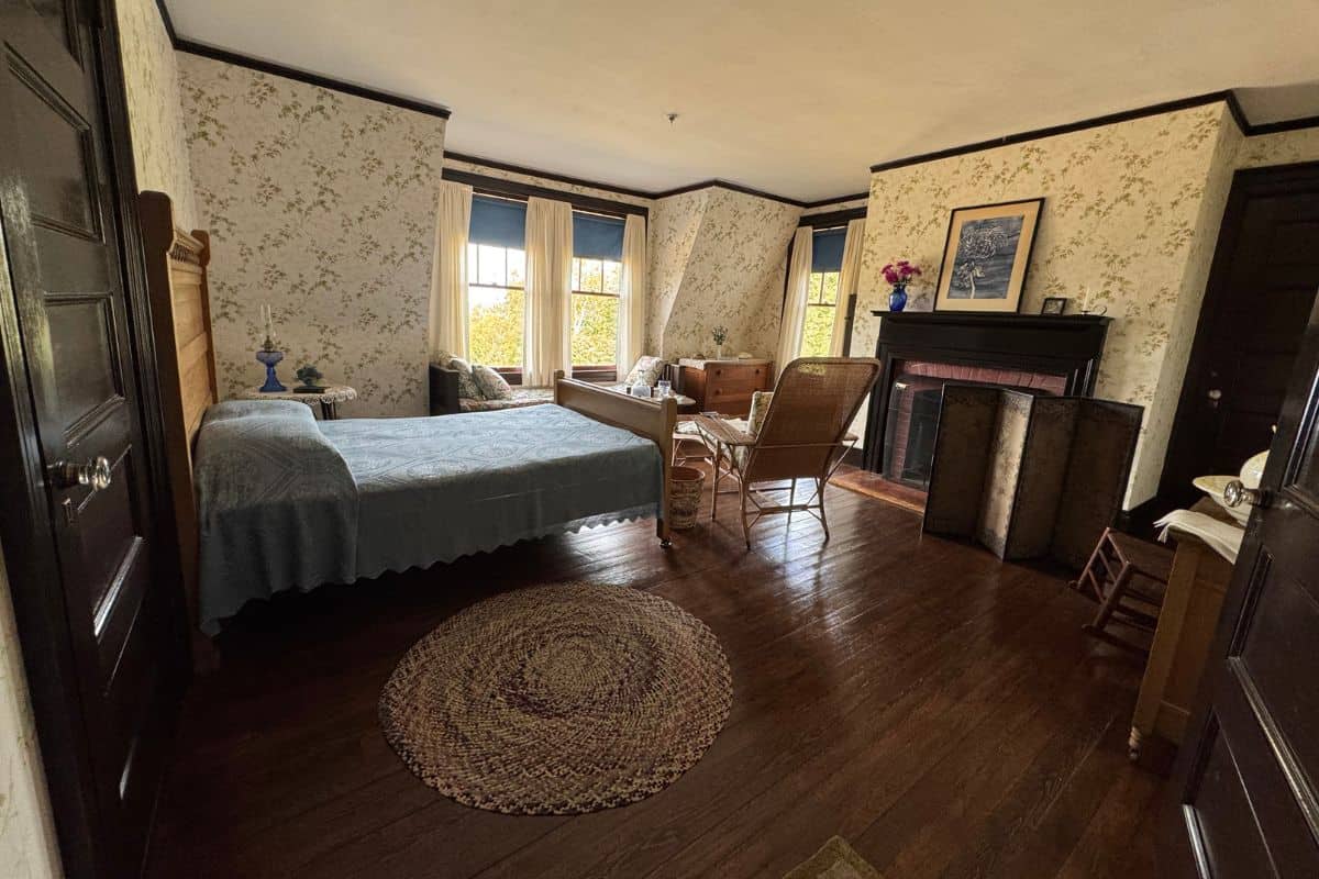 A vintage bedroom with a wooden floor, a bed with a blue cover, a rug, wicker chairs, and a fireplace evokes the charm of Lubec and nearby Campobello Island, with windows letting in soft natural light.