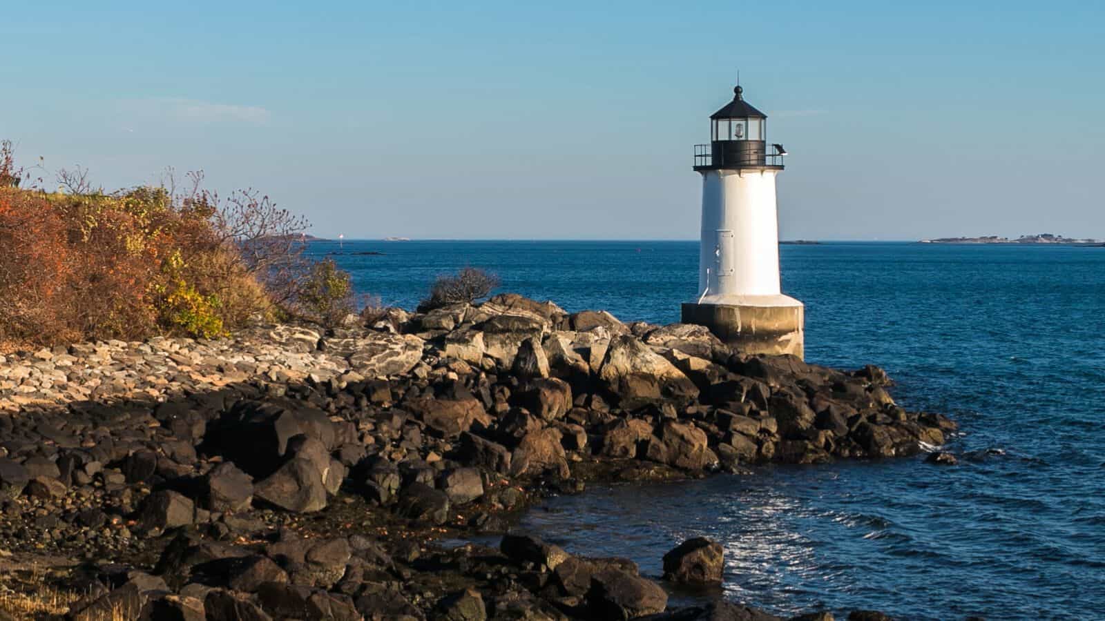 A white lighthouse stands on a rocky shoreline, surrounded by blue ocean water and sparse autumn vegetation under a clear sky.
