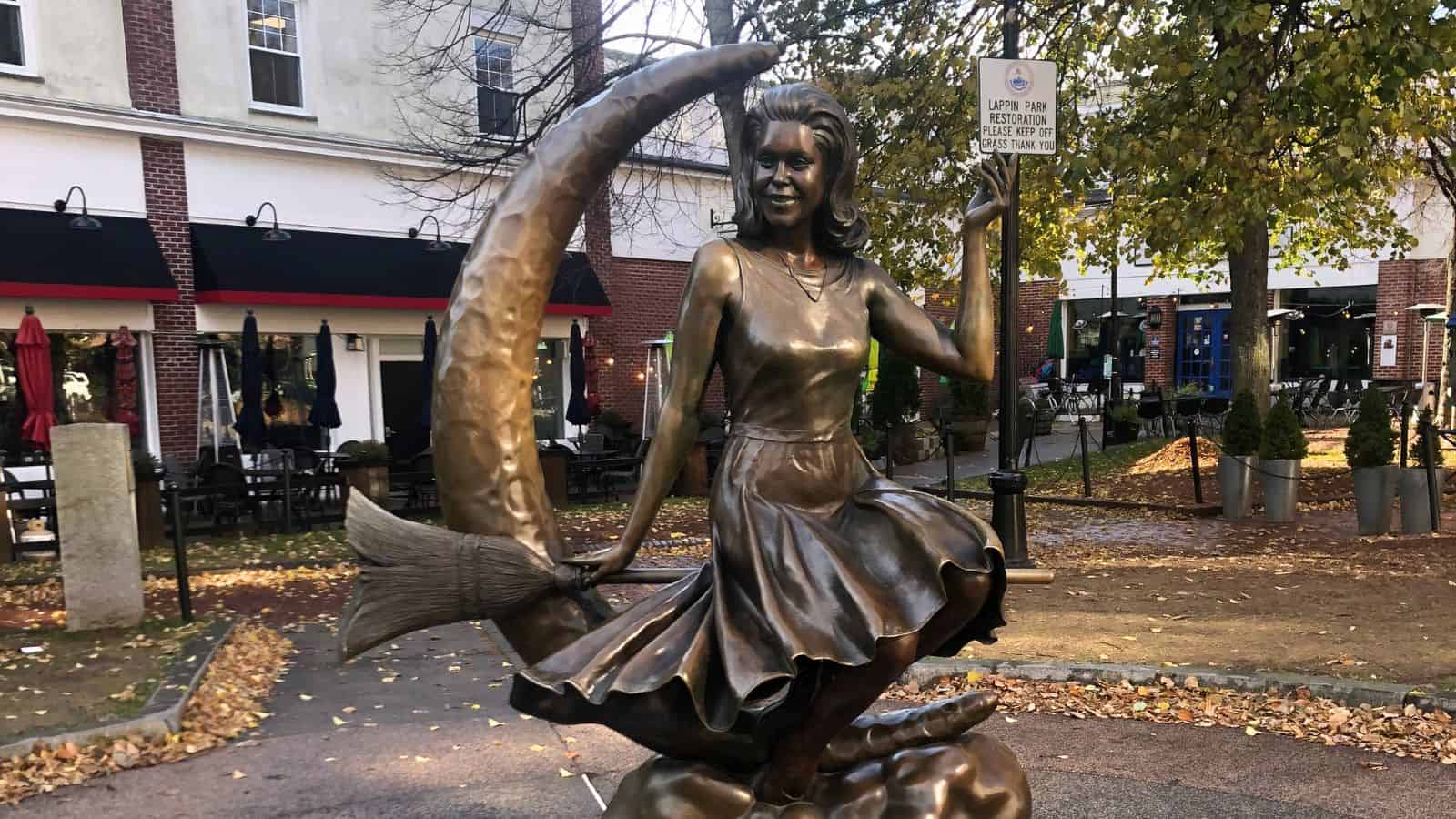Bronze statue of a woman in a dress sitting side-saddle on a crescent moon and holding a broom, located in an outdoor plaza with trees and buildings in the background.