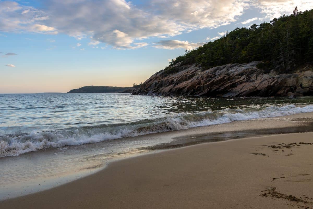 A sandy beach with gentle waves, a rocky, tree-covered hill on the right, and a partly cloudy sky at sunset.