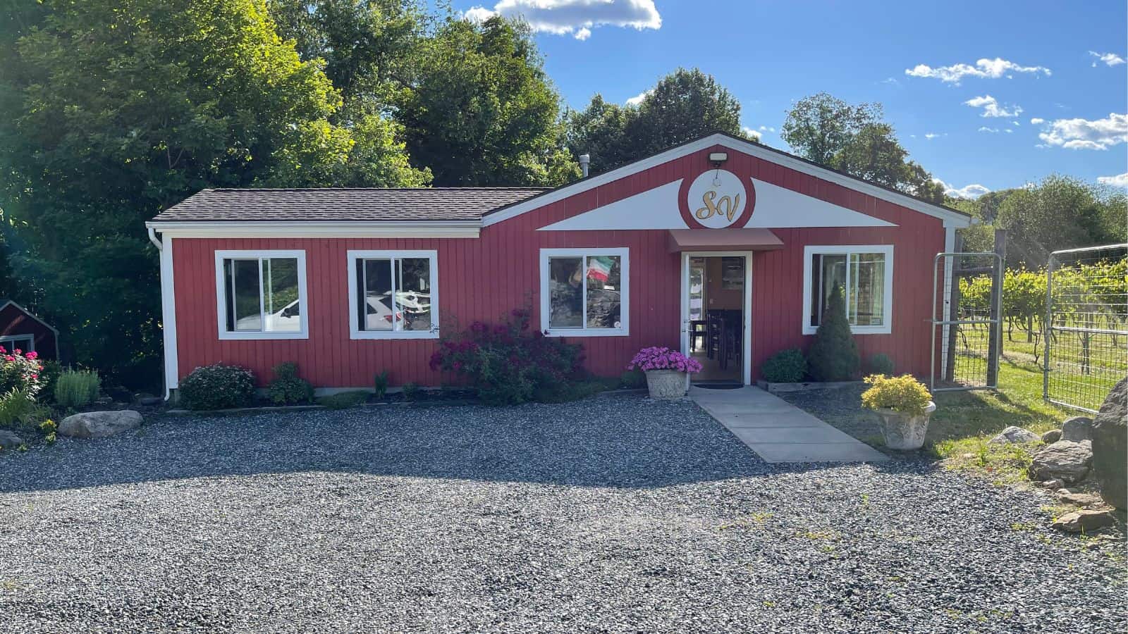 A small red building with white trim sits on a gravel lot, surrounded by trees and plants, under a blue sky with scattered clouds.