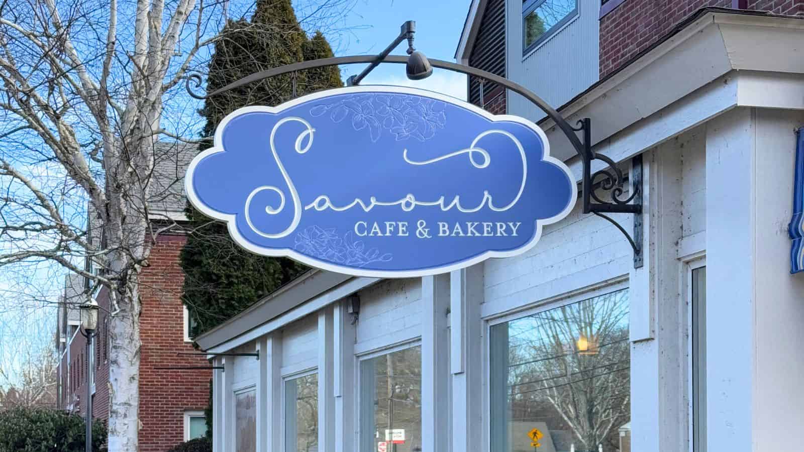 A sign reading "Savour Cafe & Bakery" hangs above the entrance of a white building with large windows and brick accents.