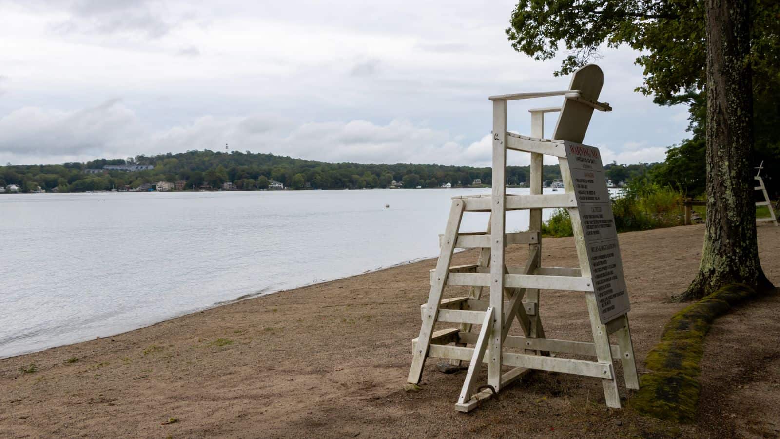 Empty lifeguard chair on a sandy beach overlooking a calm lake, with trees and houses visible across the water under a cloudy sky.