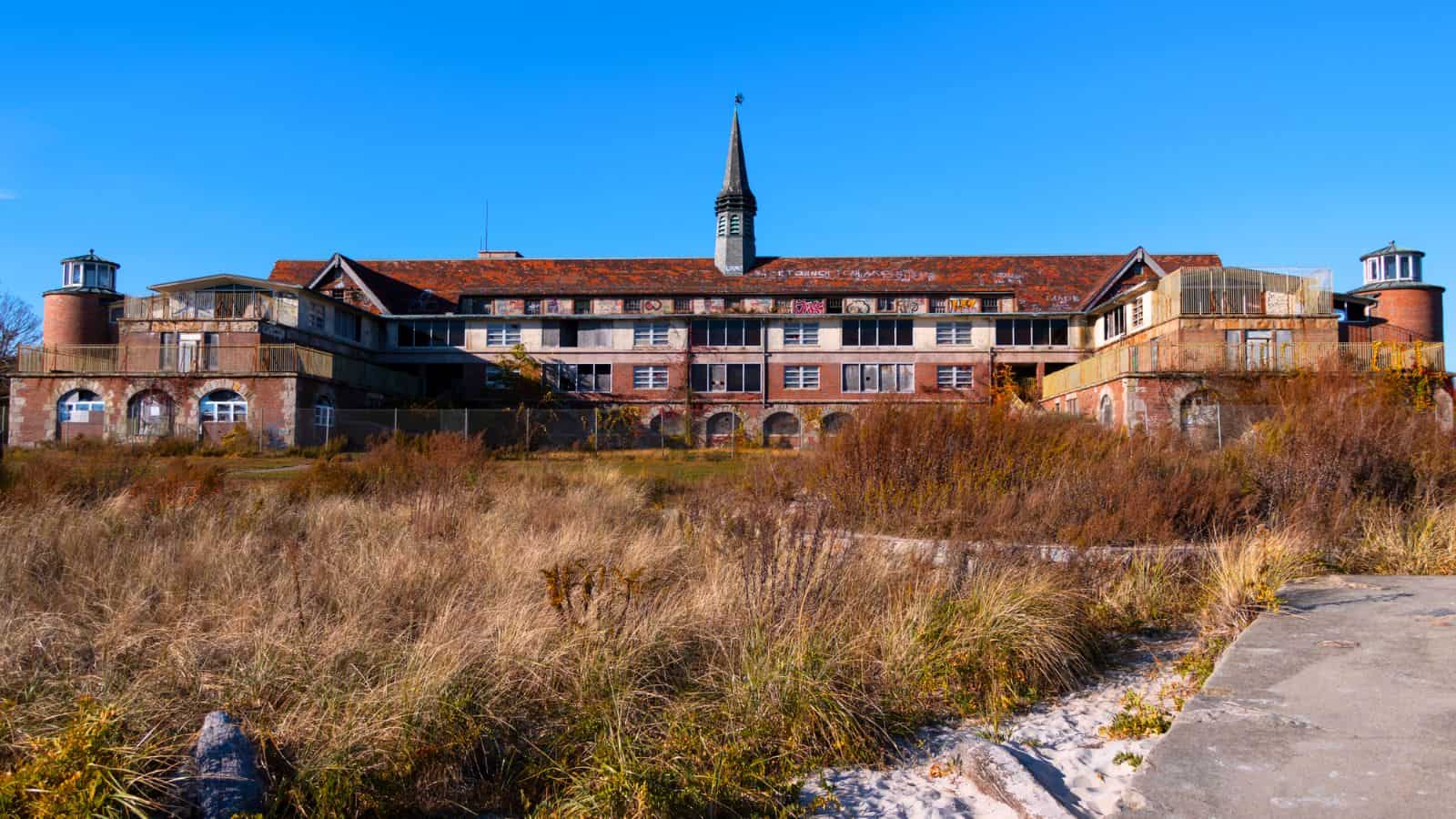 An old, abandoned brick building with a central spire and overgrown vegetation in the foreground under a clear blue sky.