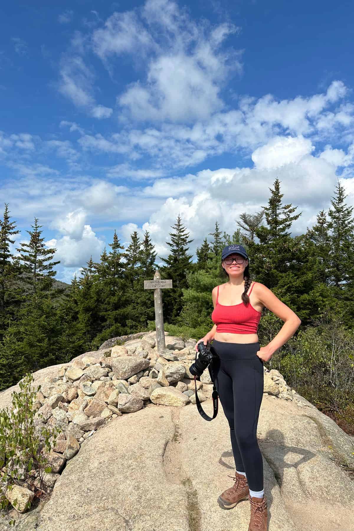 A woman in athletic wear stands on a rocky summit holding a camera, with pine trees, a wooden signpost, and a partly cloudy sky in the background.