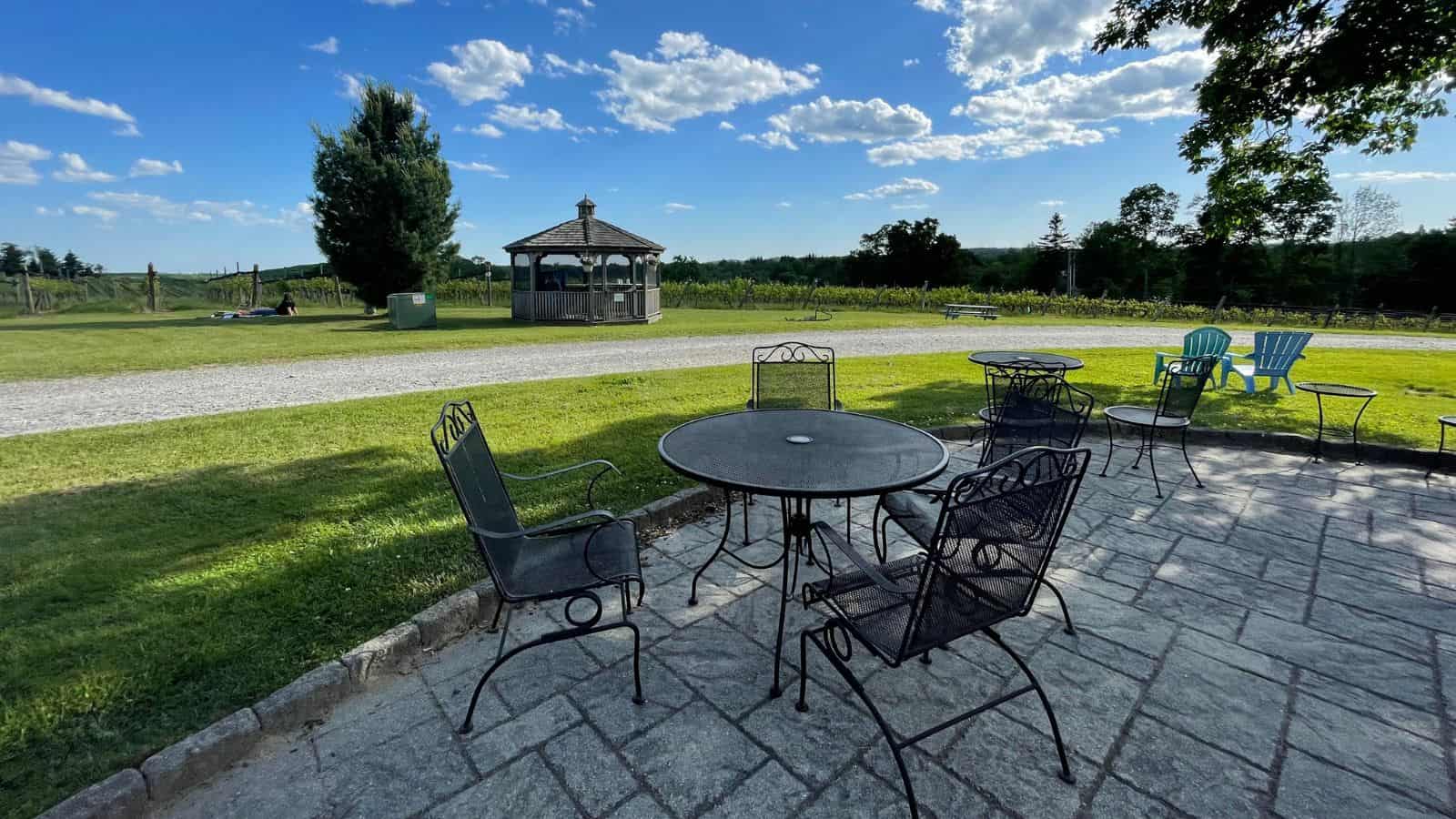 Outdoor patio with metal tables and chairs on stone pavement, facing a grassy lawn, a gazebo, and Adirondack chairs under a blue sky with scattered clouds.