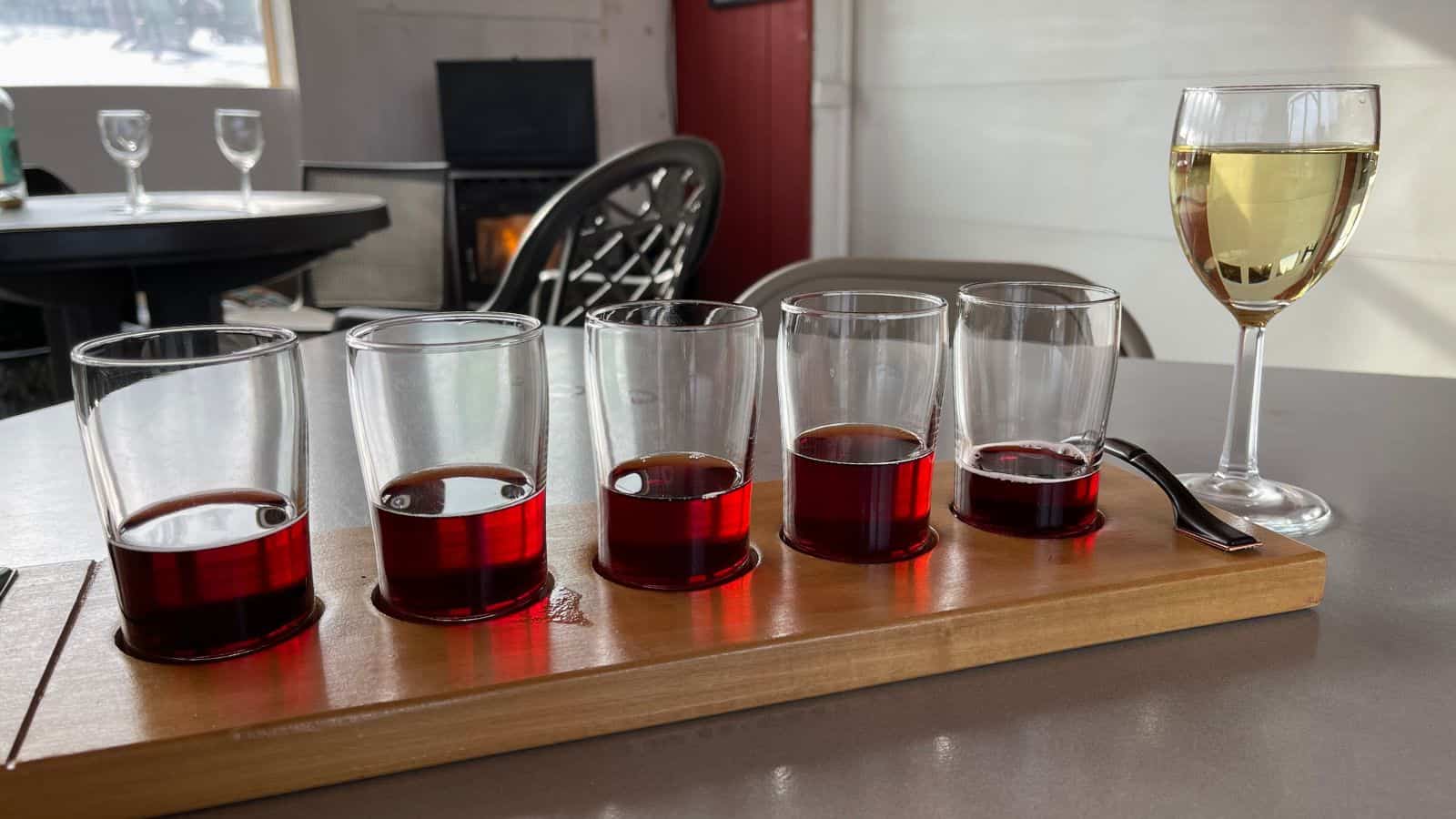 A wooden tray holds five glasses of red wine and a metal tasting spoon; a glass of white wine stands to the side on a table in a cozy indoor setting.