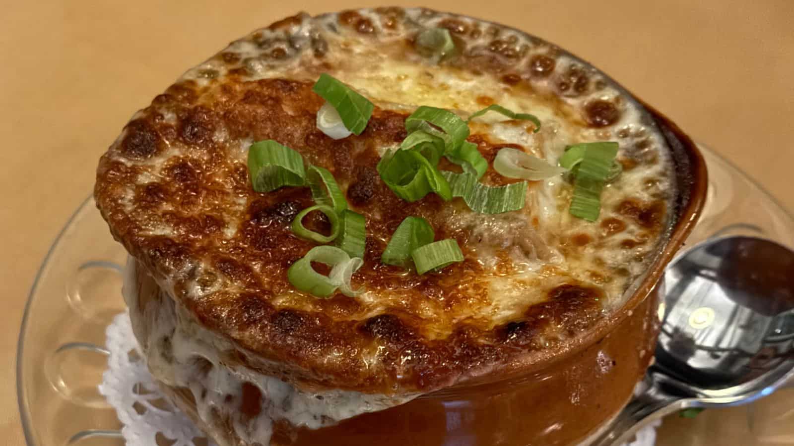 A bowl of French onion soup topped with melted cheese and sliced green onions, served on a glass plate with a spoon on the side.