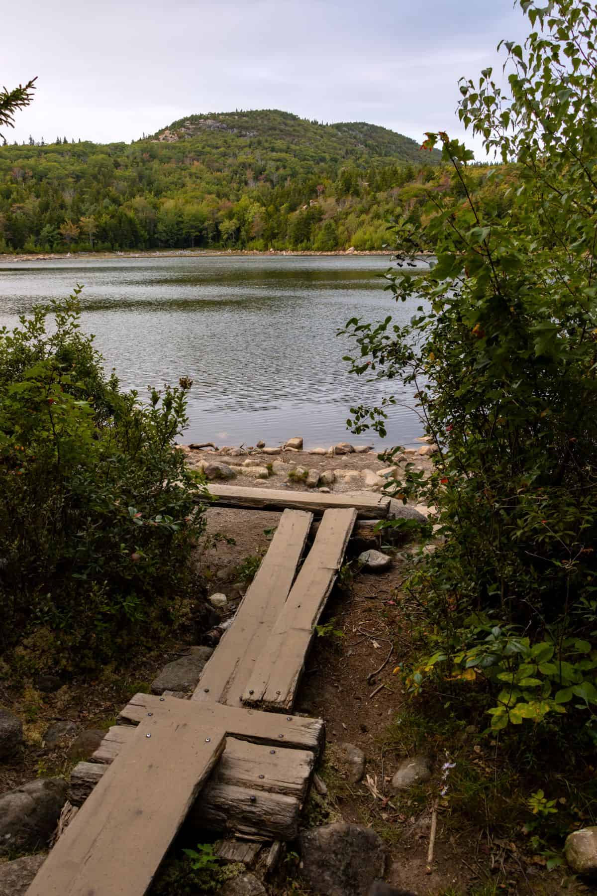 A wooden footbridge leads to a rocky lakeshore, with a calm lake and forested hills visible in the background.