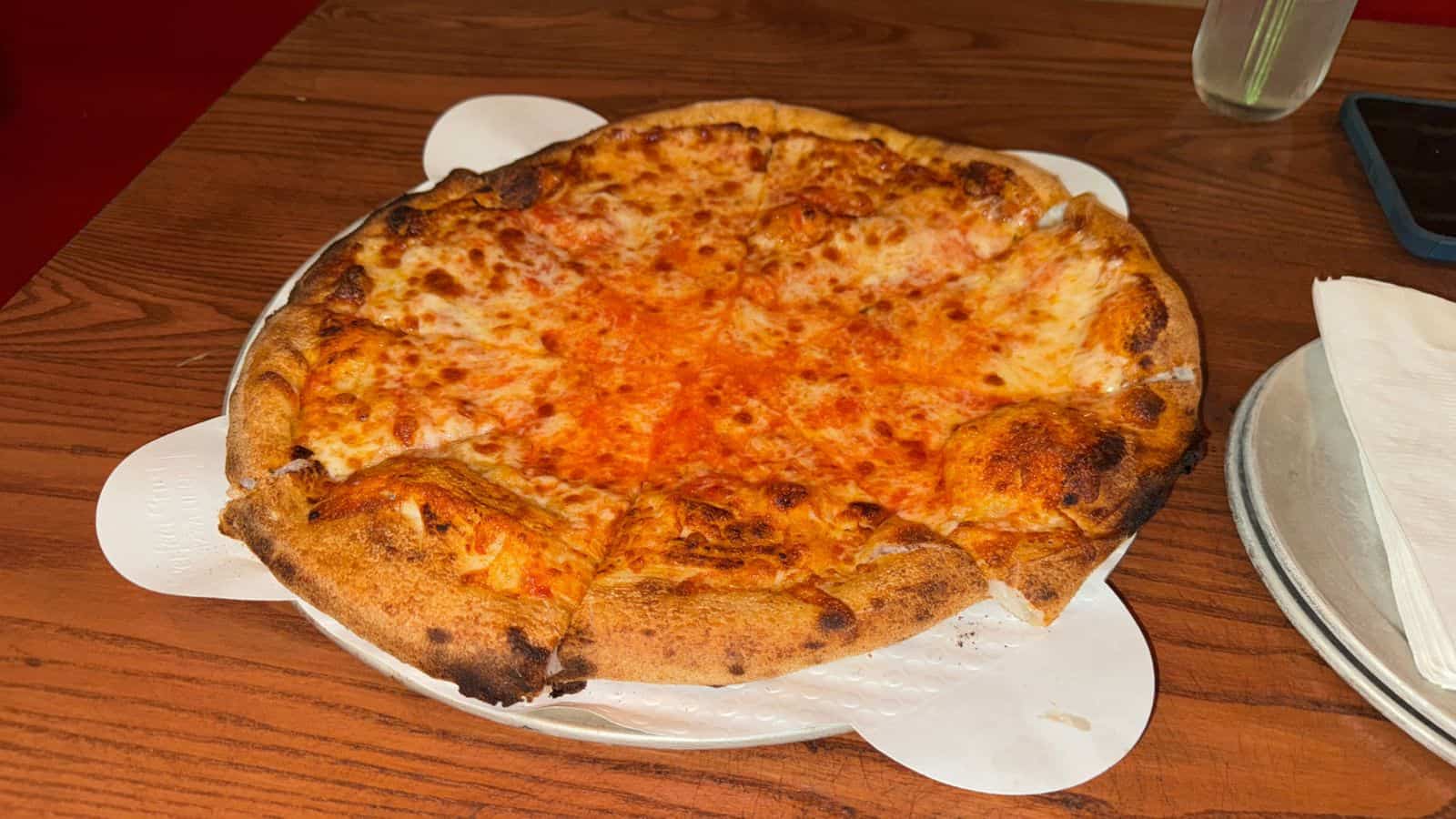 A whole cheese pizza with a thick, slightly charred crust sits on a metal tray atop a wooden table. Napkins and a smartphone are placed nearby.