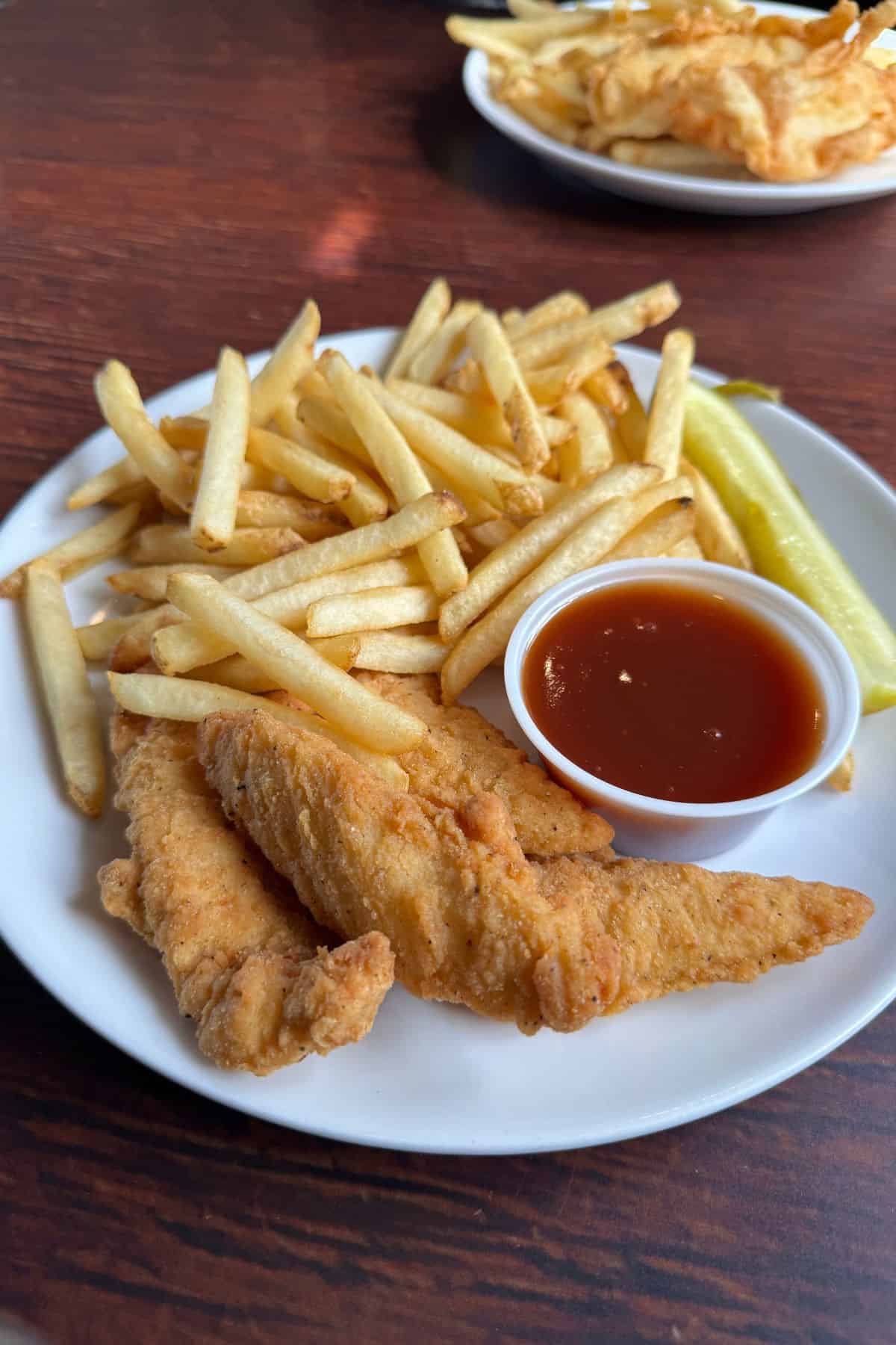 A plate of breaded fried fish fillets, French fries, a pickle spear, and a cup of dipping sauce on a wooden table. Another plate of food is in the background.
