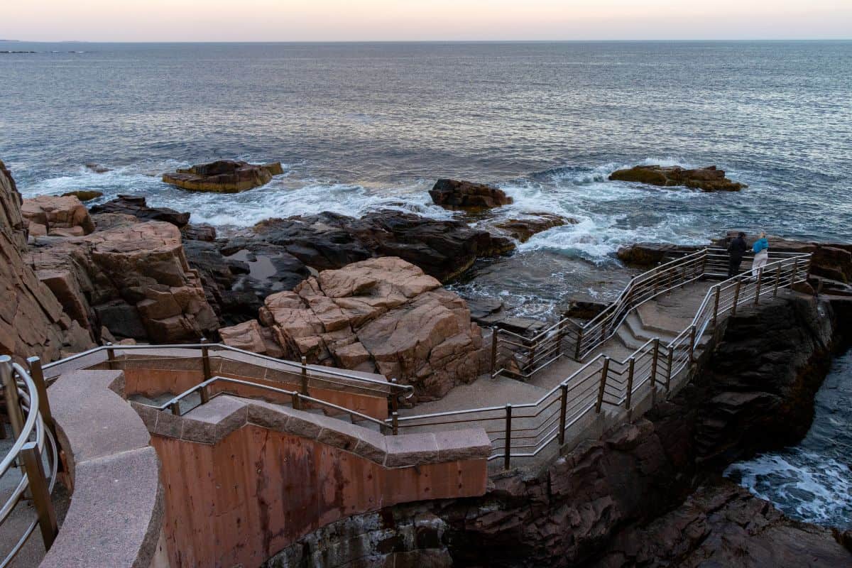 Concrete and wood stairway with railings leads down rocky cliffs to the ocean; a few people stand on the steps overlooking the water at dusk.