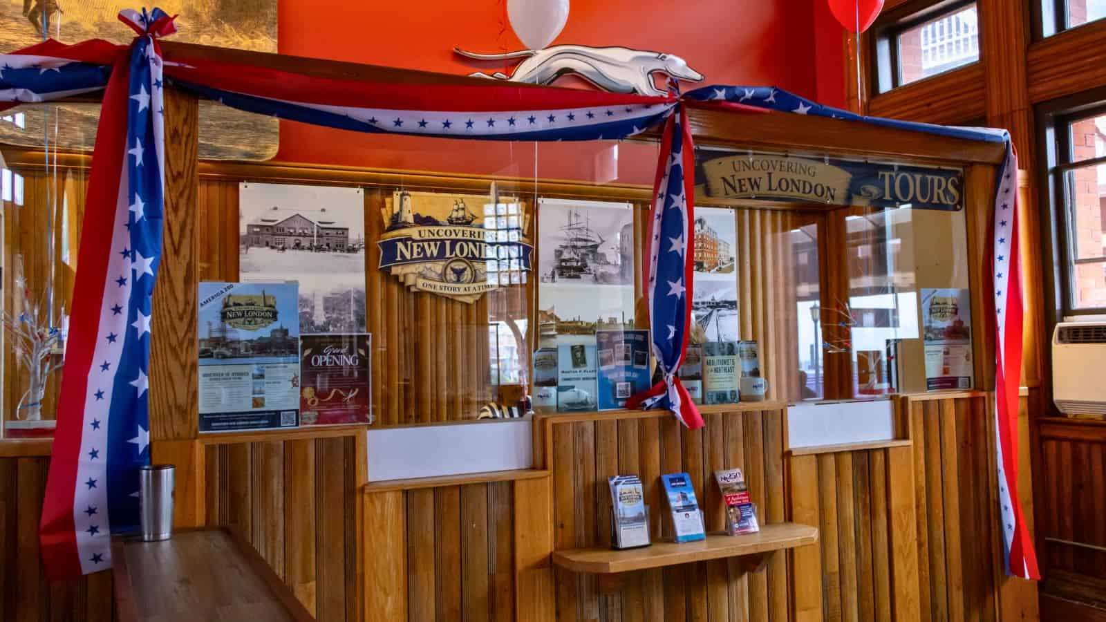 Wood-paneled ticket counter with glass windows, red, white, and blue star banners, tour brochures highlighting attractions in New London CT, and “Uncovering New London Tours” signage inside a brightly lit room.