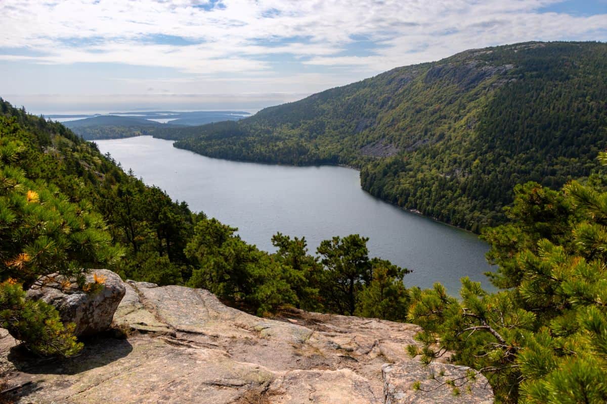 A large, calm lake is surrounded by forested hills under a partly cloudy sky, viewed from a rocky ledge with green pine trees in the foreground.
