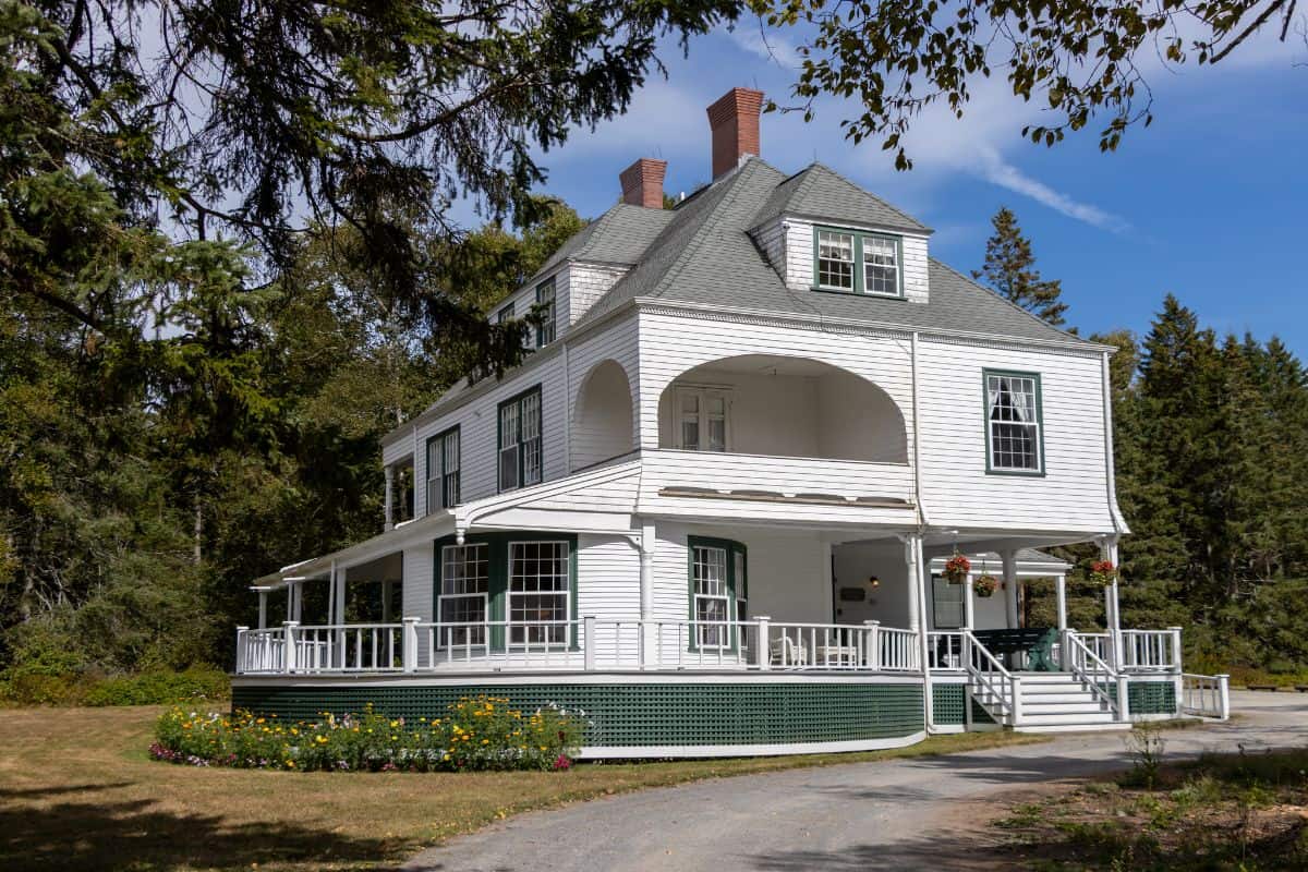 Large white two-story house with wraparound porch and green trim, set among trees with a garden and clear blue sky in the background.