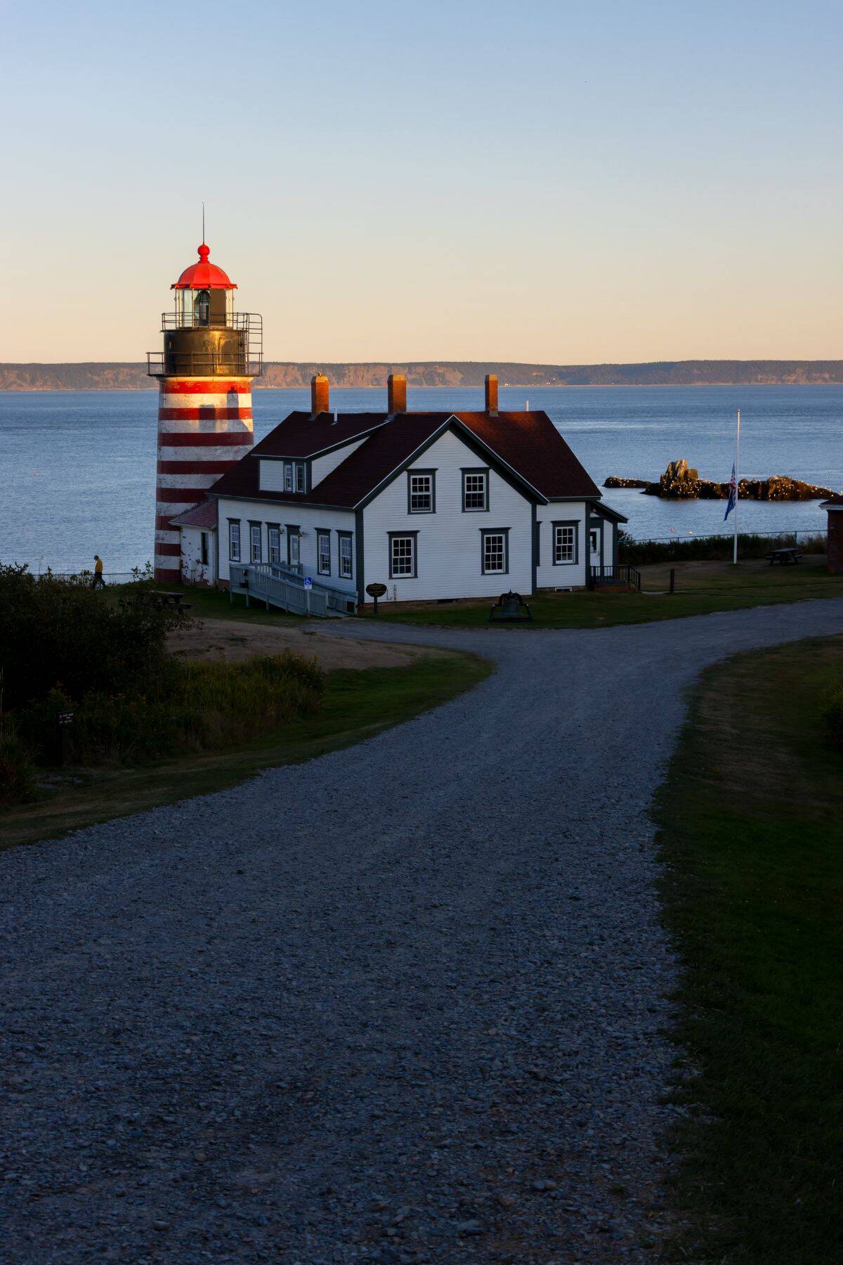 A red and white striped lighthouse with an attached white building sits near the shoreline at sunset, with a gravel road leading up to it.