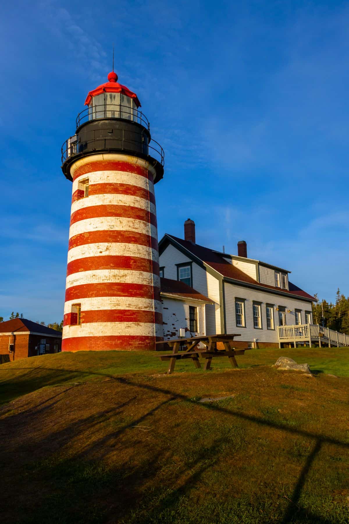 A red and white striped lighthouse stands next to a white house with a brown roof under a clear blue sky, with a picnic table in the foreground.
