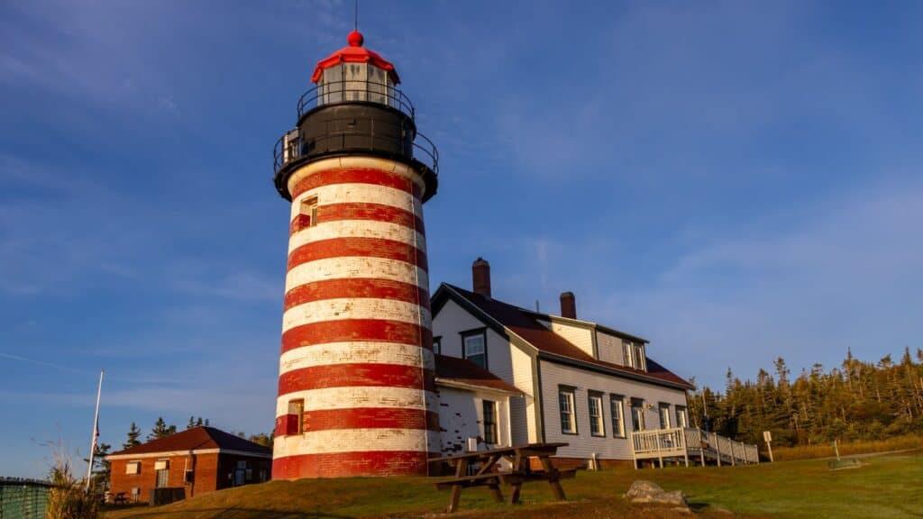 A red and white striped lighthouse stands next to a white building with a porch, surrounded by grass and trees under a clear blue sky.
