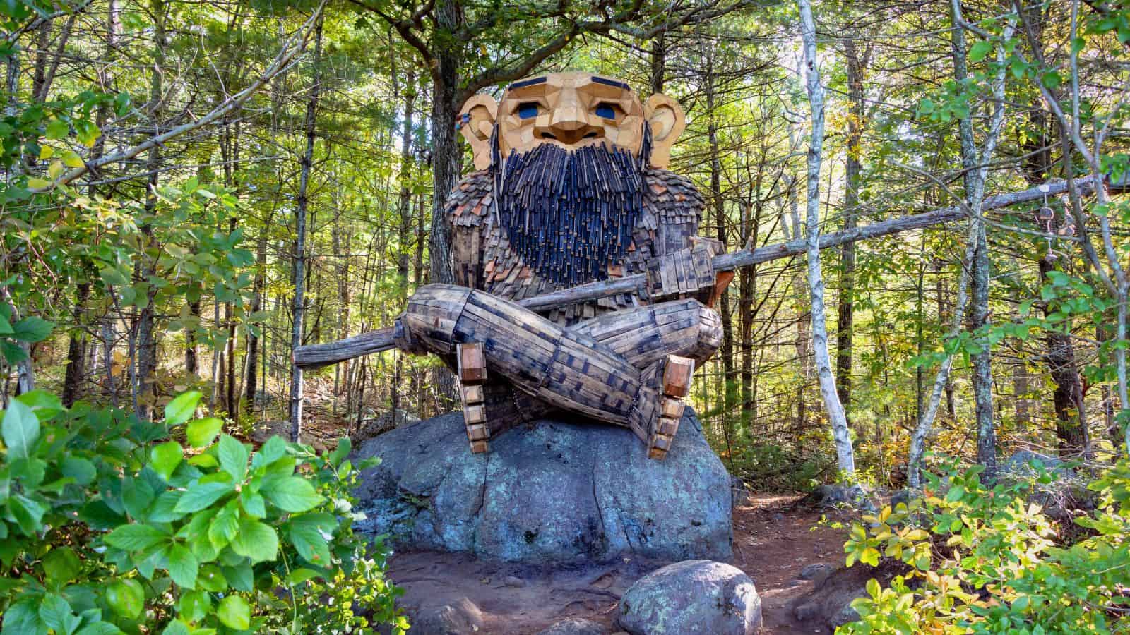 A large wooden troll sculpture sits on a rock in a forest, holding a long stick across its lap, surrounded by green trees and foliage.