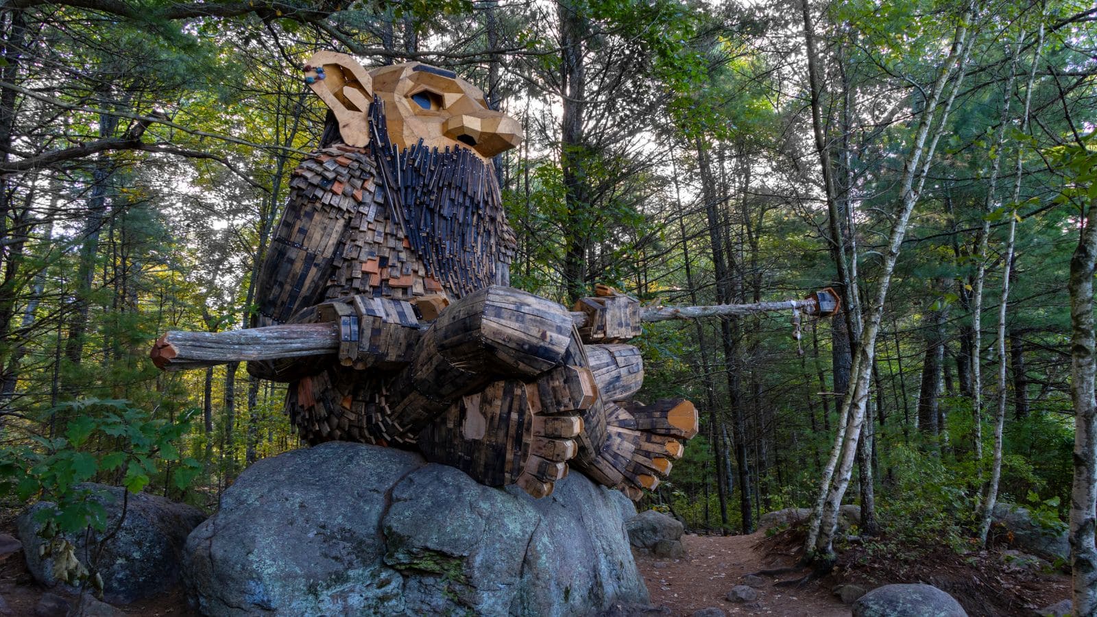 A large wooden troll sculpture sits on a boulder in a forested area, holding a long staff made of tree branches.