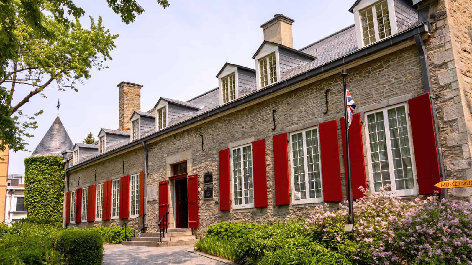 A stone building with red shutters and multiple dormer windows, surrounded by greenery and flowering bushes, under a clear sky.
