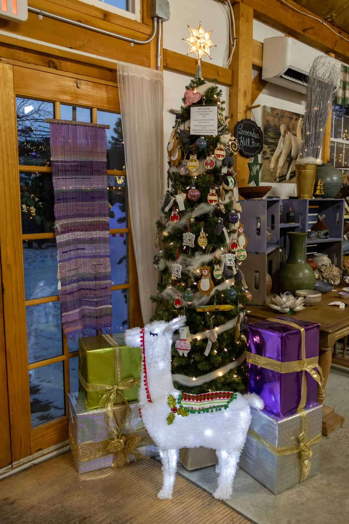 A decorated Christmas tree with assorted ornaments, a white llama figure reminiscent of Lavender Hill Farm Alpacas, and large wrapped presents stands indoors near wooden doors and woven textiles.