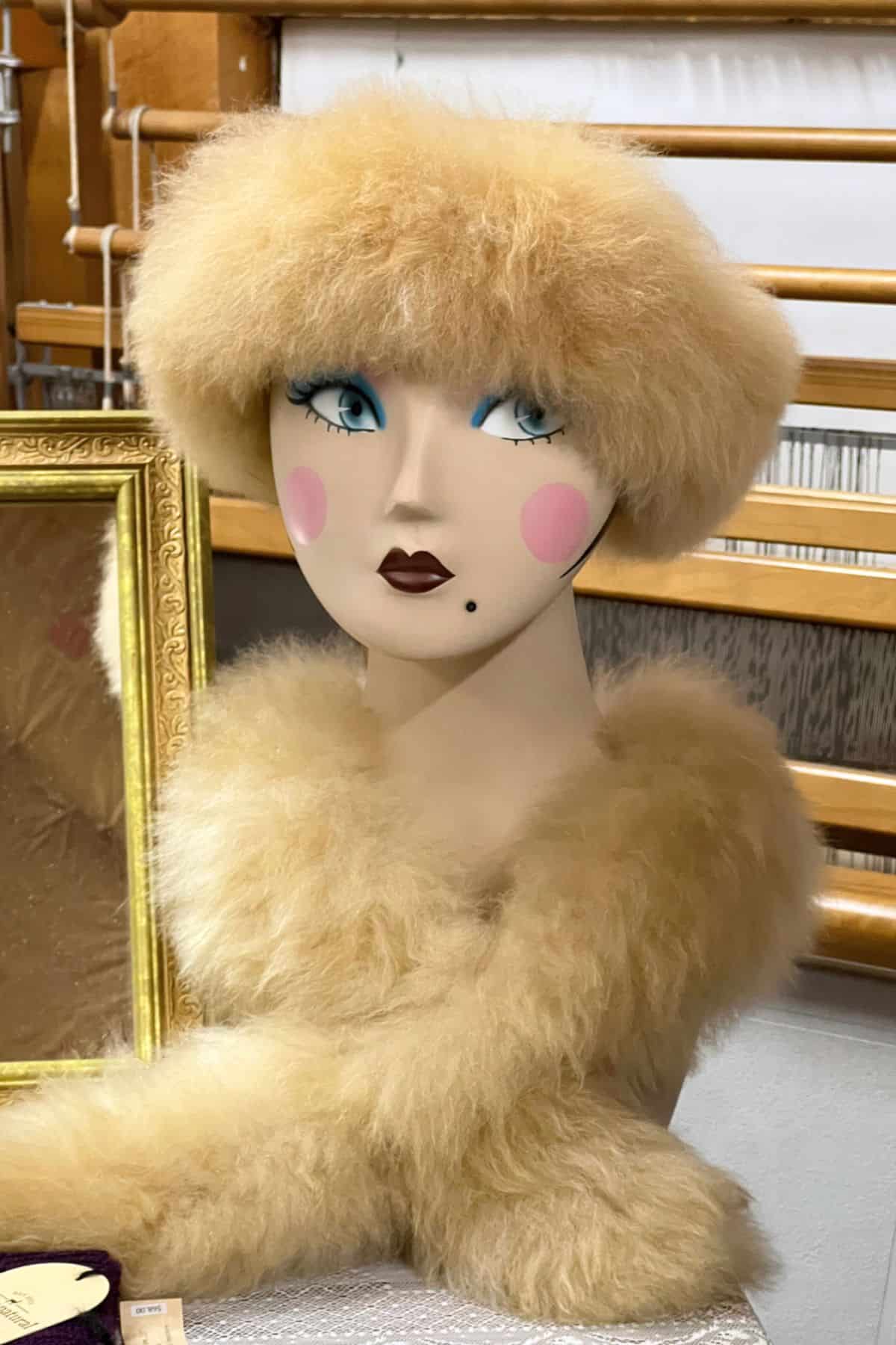 A mannequin head with painted facial features and pink blush, wearing a fluffy, light brown fur hat and matching scarf made from Lavender Hill Farm Alpacas fiber, displayed indoors next to a gold-framed mirror.