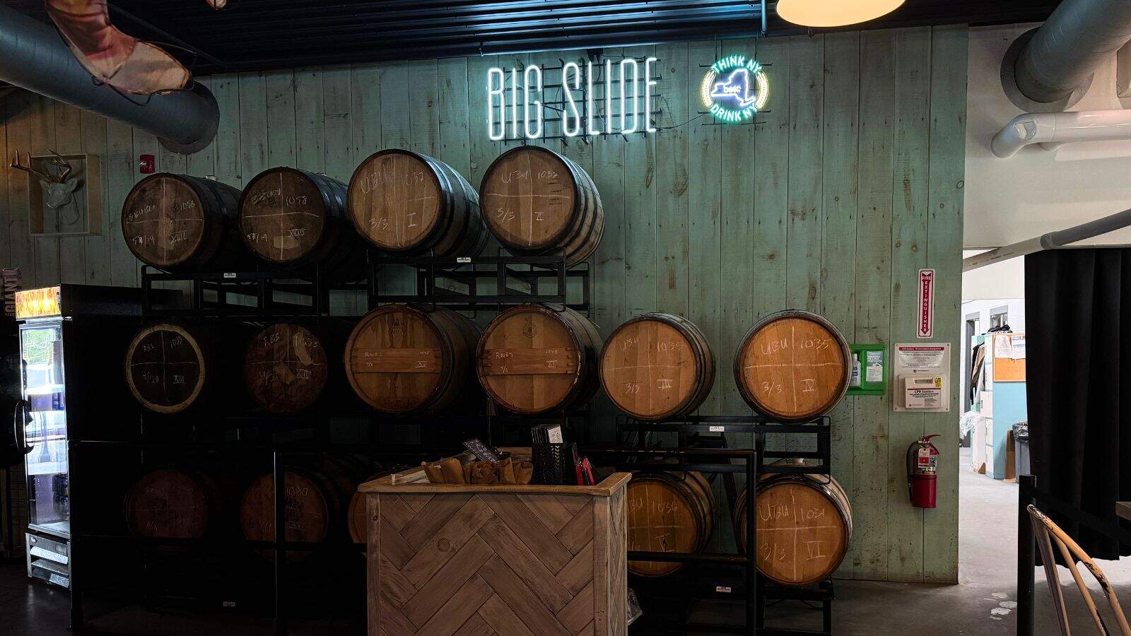A stack of wooden barrels on black racks sits against a green wooden wall with a neon "Big Slide" sign above in an indoor brewery setting.