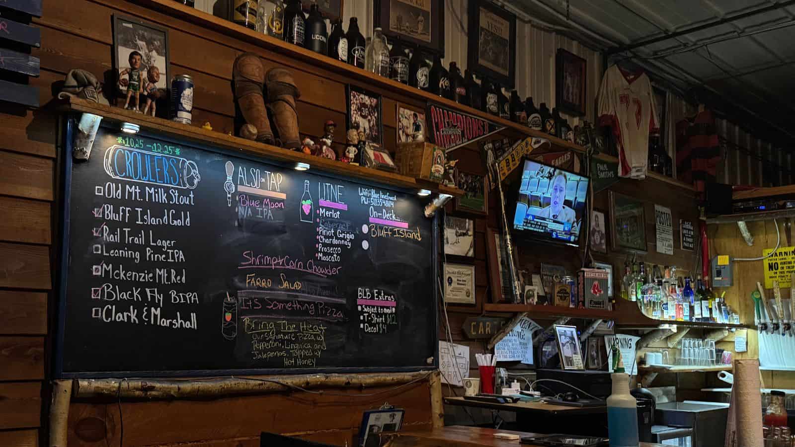 Bar interior with a chalkboard beer menu, liquor bottles on shelves, framed photos on wood walls, a hanging TV, and various decorations behind the counter.