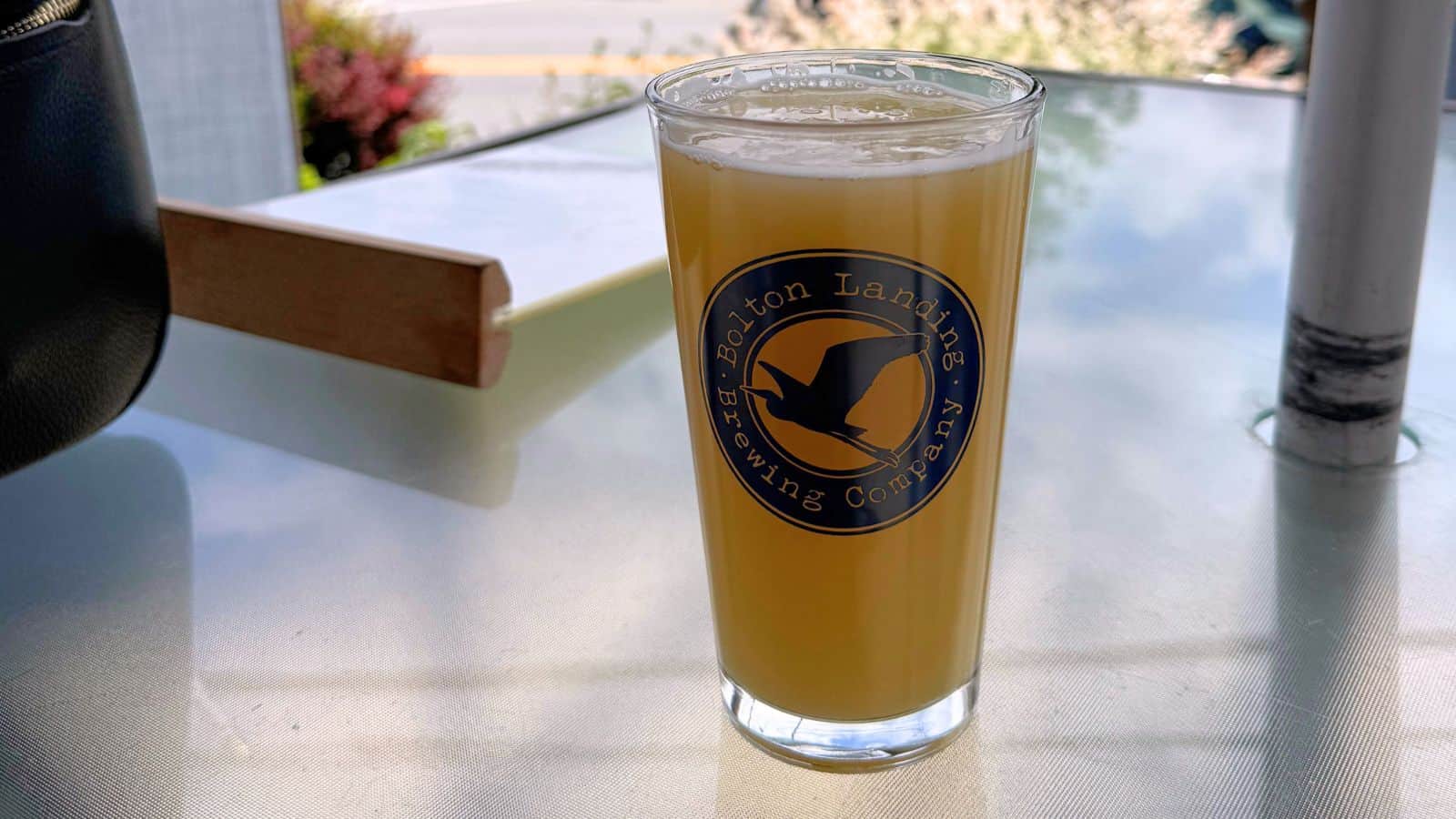 A pint glass filled with hazy beer sits on a glass table. The glass displays the Acton Landing Brewing Company logo. Background includes a blurred outdoor scene.