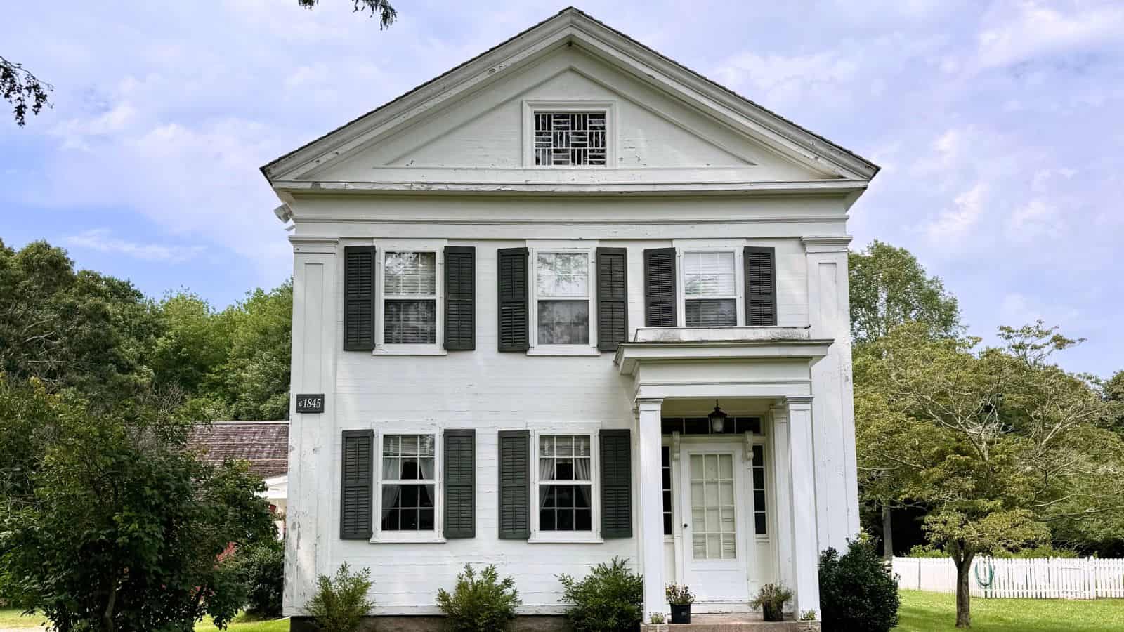 A white two-story house with green shutters, a gabled roof, and a small front porch, surrounded by grass and trees under a partly cloudy sky.