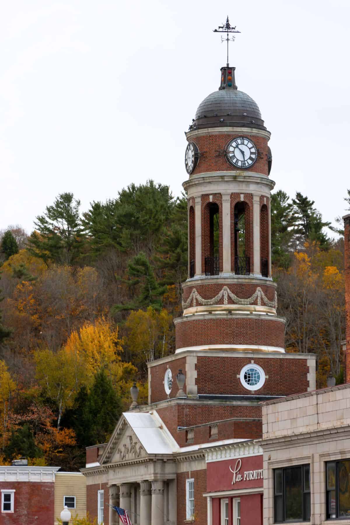 Brick clock tower with a dome and weathervane rises above Saranac Lake, surrounded by autumn trees and nearby brick buildings under a cloudy sky.