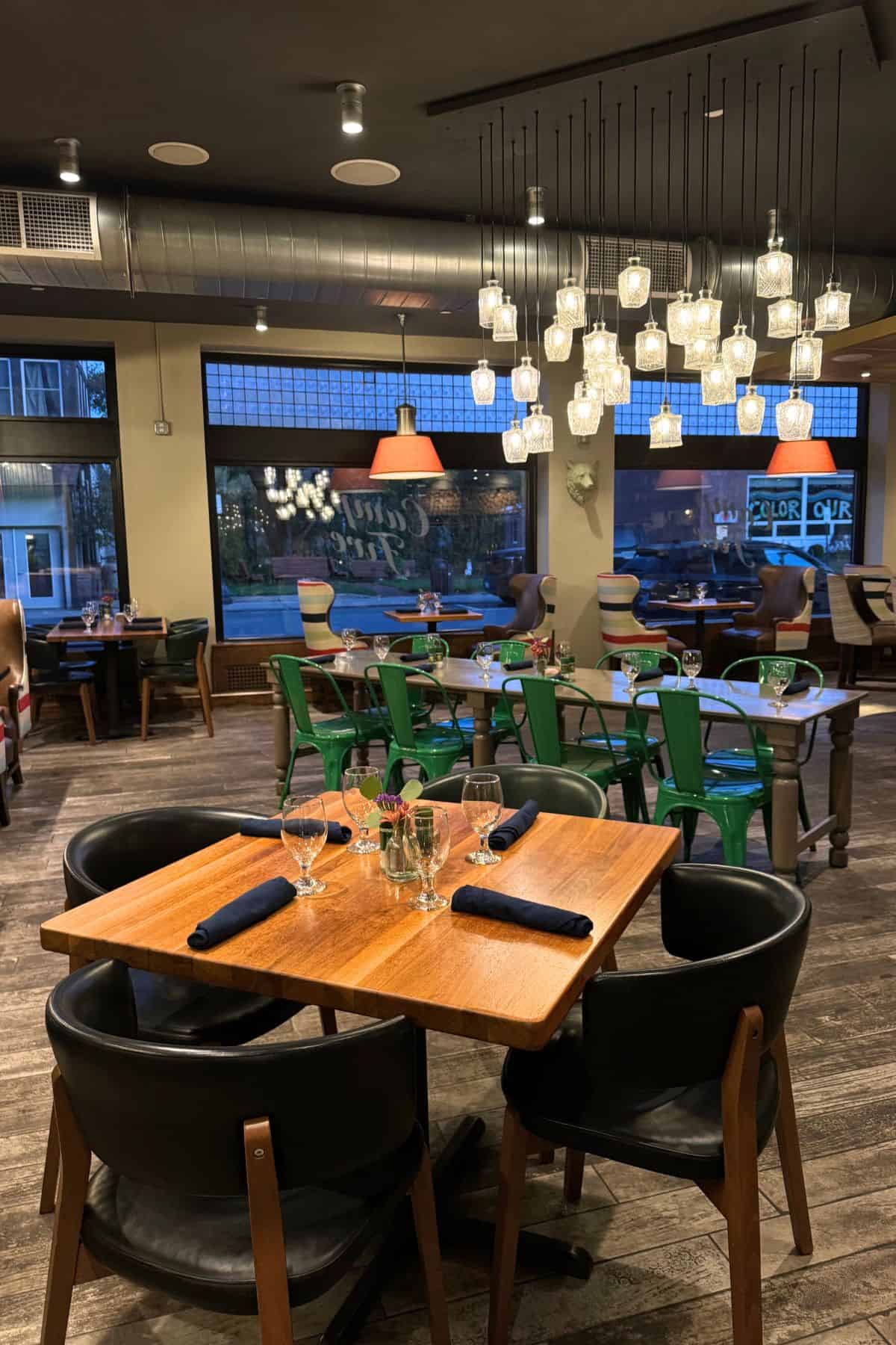Modern restaurant interior with wooden tables, black chairs, green metal chairs, glassware, napkins, large windows, and hanging glass pendant lights.