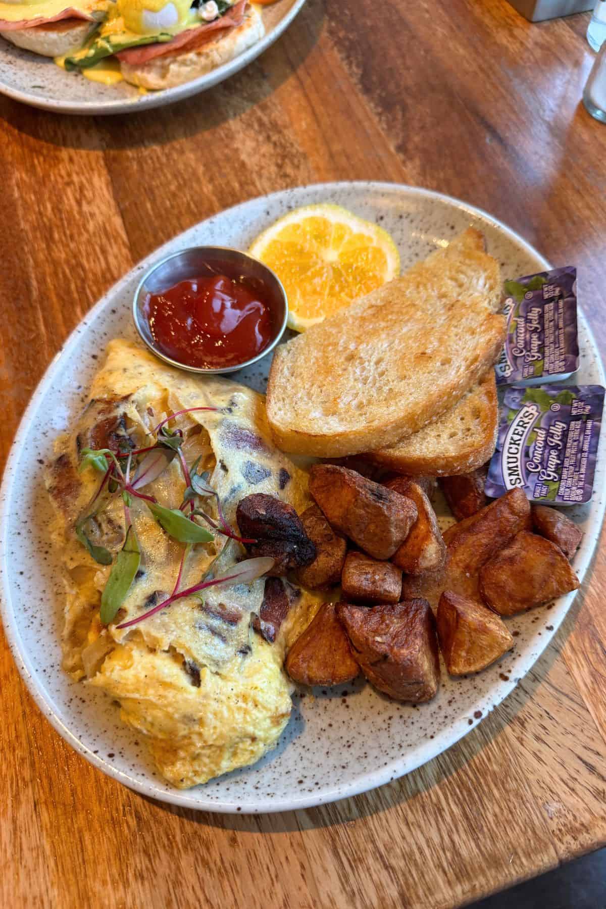 A breakfast plate in Saranac Lake features an omelette, roasted potatoes, toast with grape jelly, orange slices, and a metal cup of ketchup on a speckled dish, all set on a wooden table.